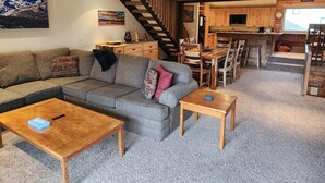 Un salon chaleureux avec un canapé d’angle, une table basse en bois et un escalier menant à une cuisine.