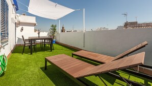 Een terras op het dak met kunstgras, houten lounge fauteuils, een tafel met stoelen en een witte tafelkleed.