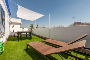 Terraço no telhado com grama sintética, espreguiçadeiras de madeira, uma mesa com cadeiras e um tecido branco.