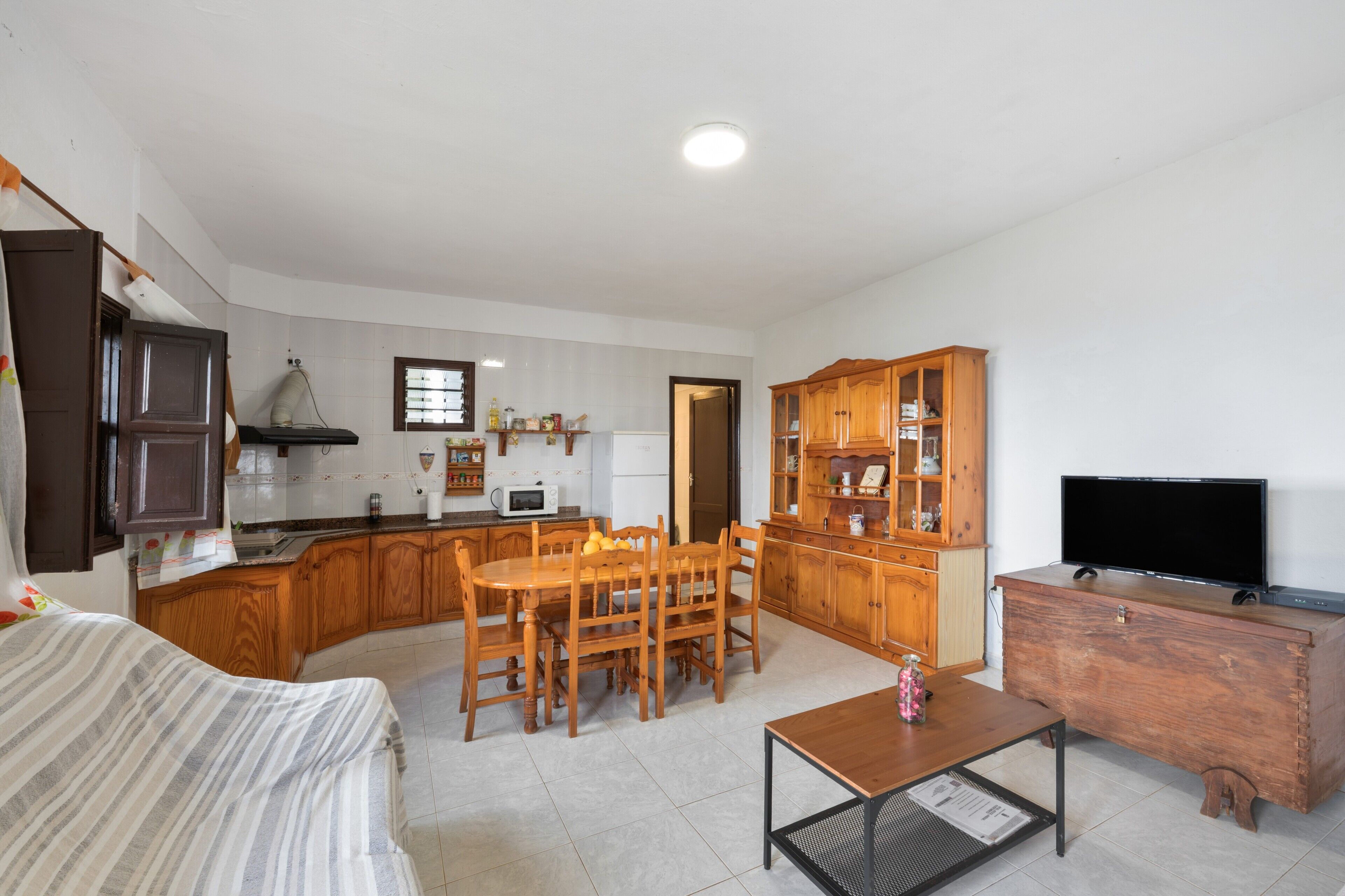 A compact kitchen with wooden cabinets, a dining table with chairs, a television on a wooden dresser, and a bed with striped bedding.