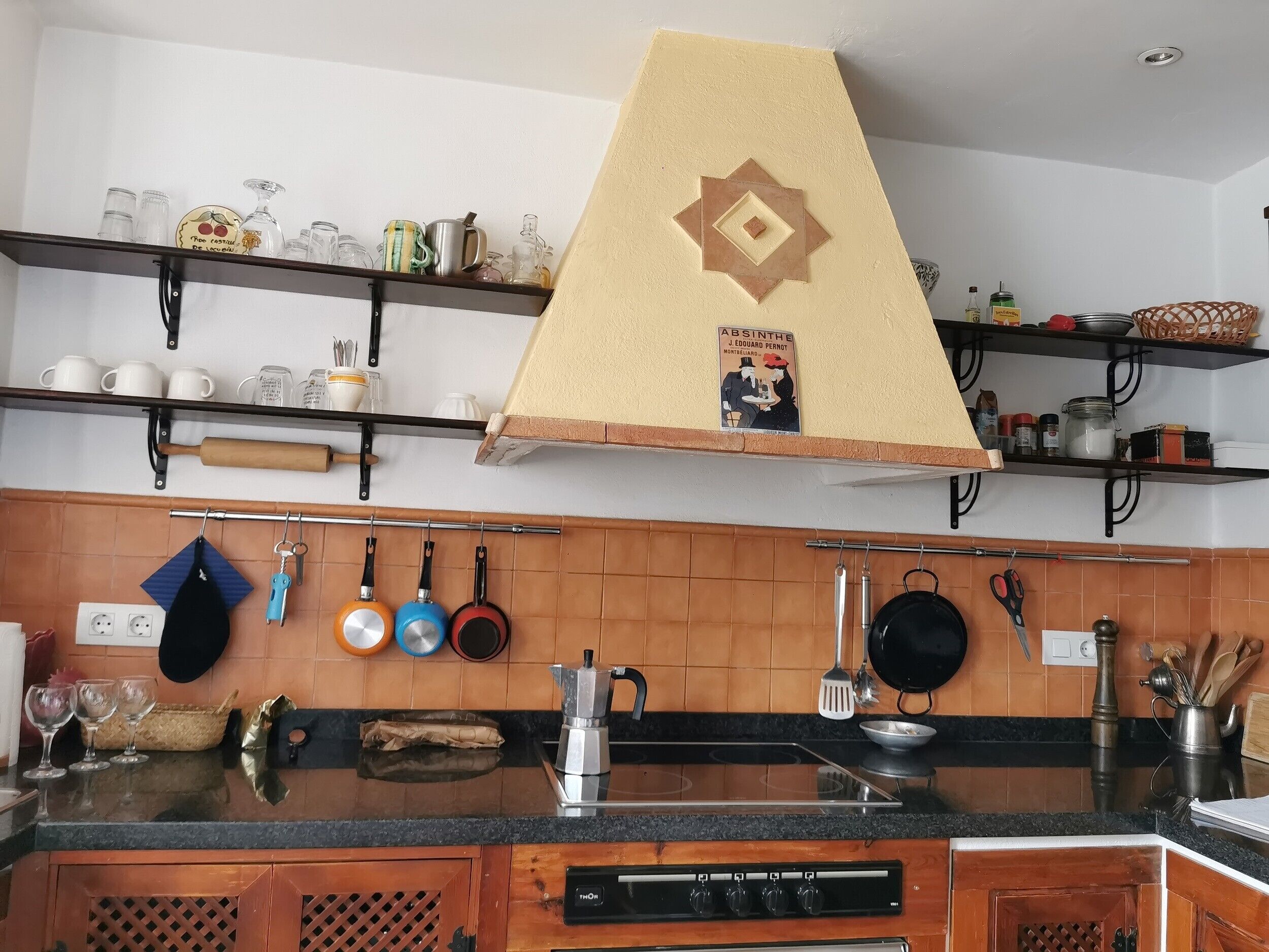 A kitchen with a central island, a stove, hanging utensils, and various kitchenware on shelves.