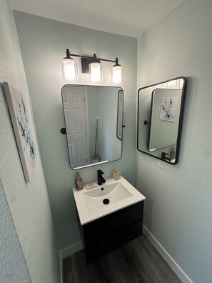 Un baño con lavamanos blanco, tocador negro y dos espejos con luces empotradas.