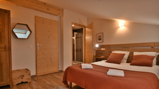 A bedroom with a wooden headboard, a large bed with red bedding, a wooden wardrobe, and a hexagonal mirror on the wall. - Les Primeveres - Type Loft 10p (Saint-Jean-de-Sixt)