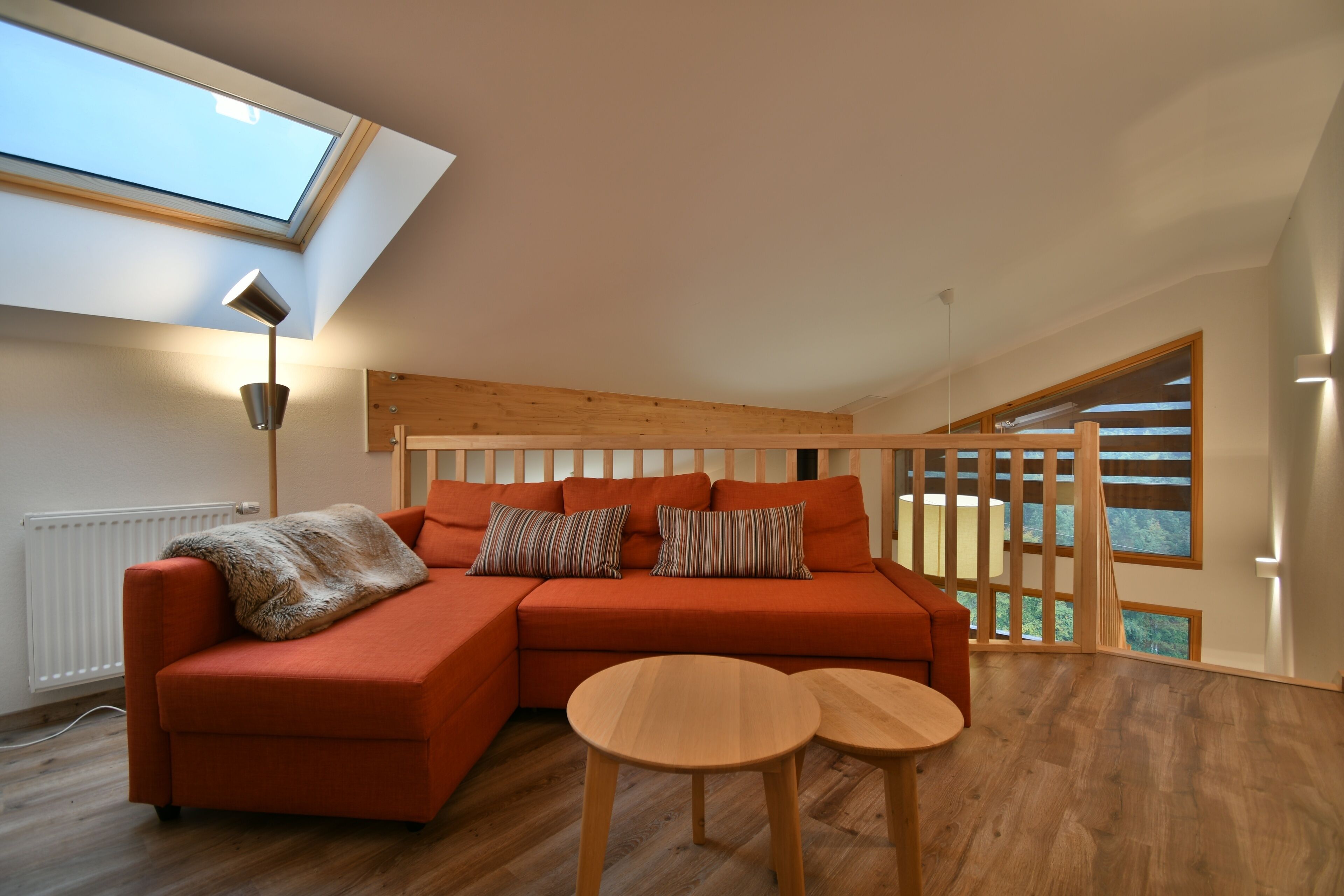 A modern living room with a sectional sofa, two round wooden tables, a skylight, and a large window with a view.