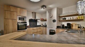 A modern kitchen with wooden cabinets, stainless steel appliances, a large island with a sink, and shelves with glassware and bottles. - Les Primeveres - Type Loft 10p (Saint-Jean-de-Sixt)