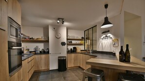 A modern kitchen with wooden cabinets, a stainless steel oven, a coffee maker, and a bar area with stools. - Les Primeveres - Type Loft 10p (Saint-Jean-de-Sixt)