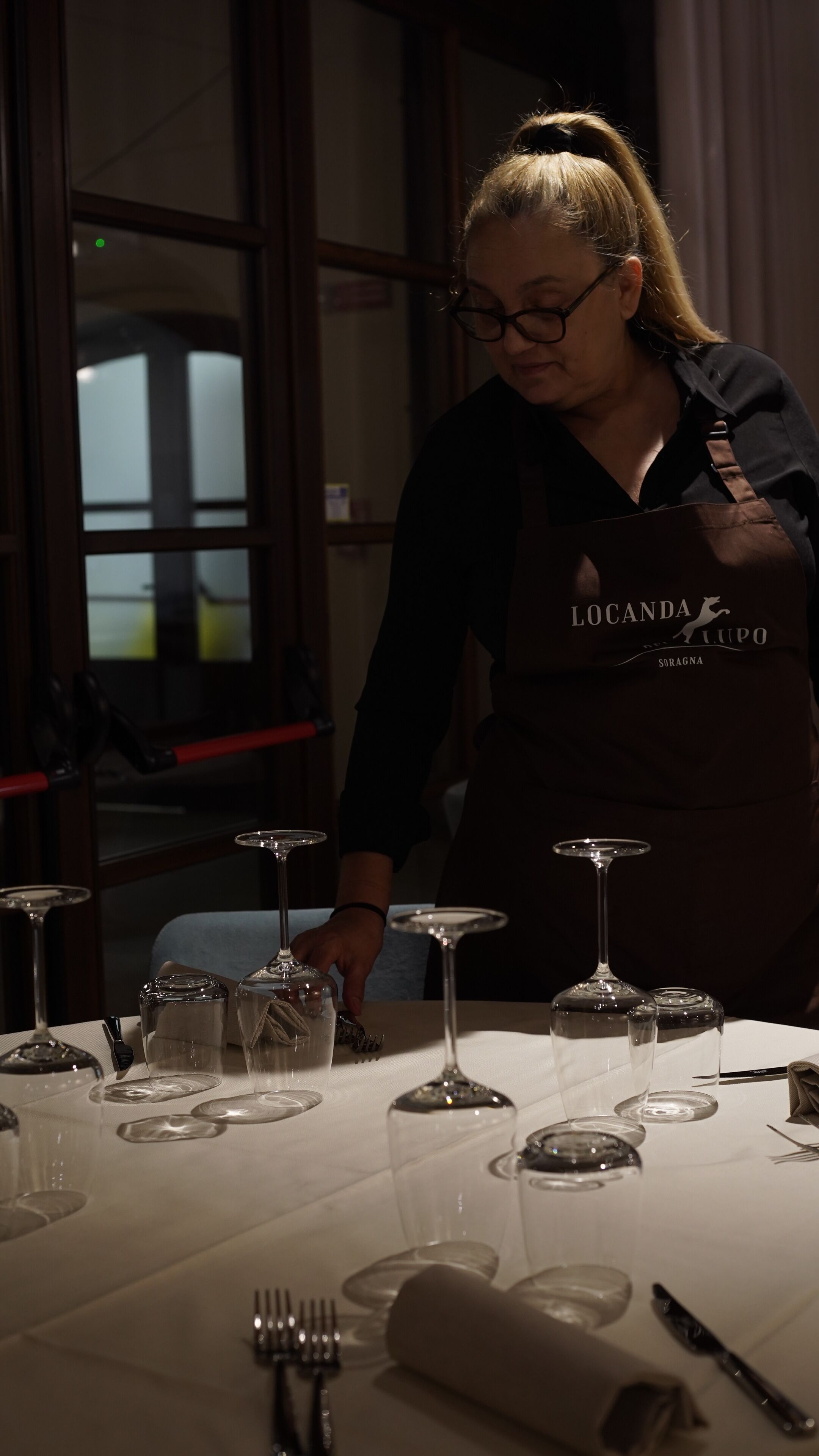 Une personne portant un tablier foncé, sur lequel on peut lire le texte « Iocanda Luipo Soragna », est en train de disposer des verres à vin sur une table recouverte d’une nappe blanche.