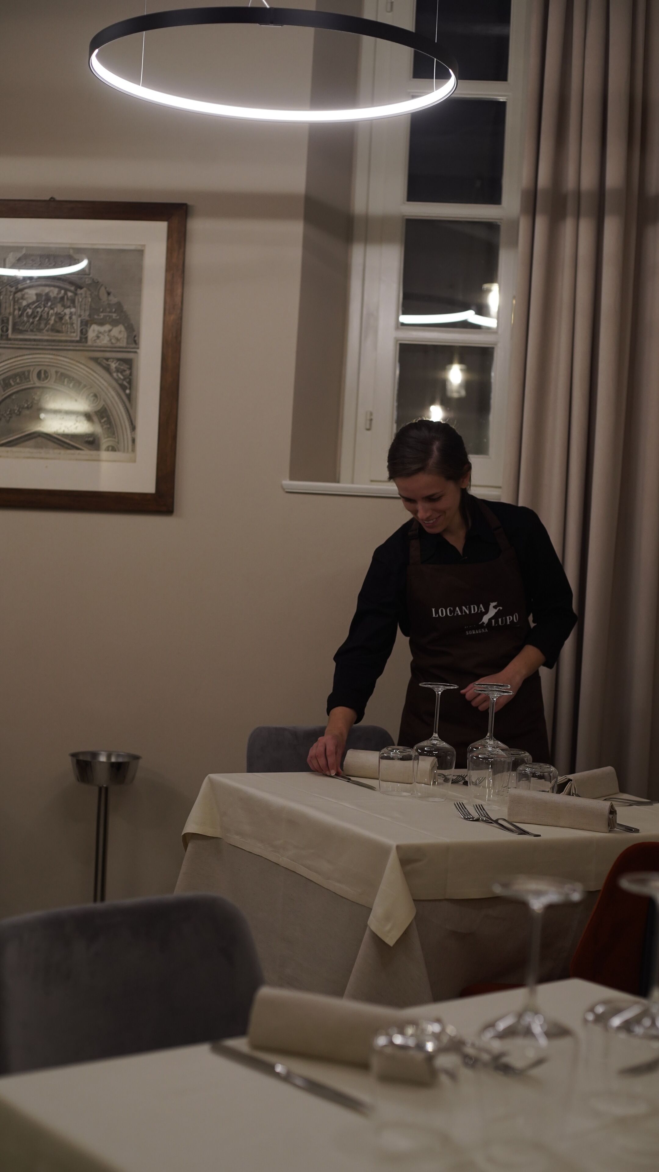 A dining area with a table set for a meal, a waiter in an apron, and a framed picture on the wall.