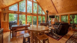 Una cabaña espaciosa con techo alto, ventanas grandes, comedor, sala de estar con chimenea y vistas al bosque.