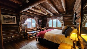 Un dormitorio rústico de cabaña de madera con una cama grande, paredes de madera, un banco y una ventana con cortinas.