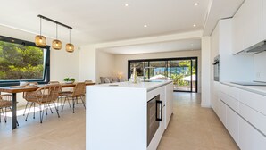 Een moderne keuken met een centraal eiland, een eethoek met rieten stoelen, en een groot raam met uitzicht op groen.