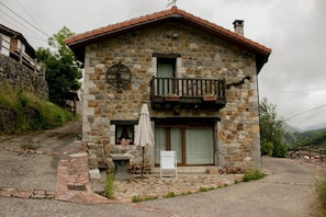 A stone house with a balcony, a small outdoor seating area, and a signboard. - Llar de Cosme: Casa rural en el corazón del Parque Natural de Redes, Asturias (Caso)