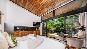A modern bedroom with a large bed, a TV, wooden ceiling, and a view of a lush garden through large windows.