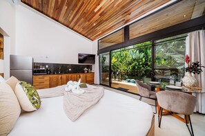 Ein modernes Schlafzimmer mit einem großen Bett, einem Fernseher, Holzboden und Blick auf einen üppigen Garten durch große Fenster.