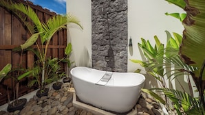A freestanding bathtub in a garden setting with a wooden fence, stone wall, and various tropical plants.