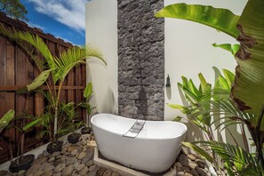 Eine freistehende Badewanne in einer Gartenanlage mit Holzzaun, Steinmauer und verschiedenen tropischen Pflanzen.