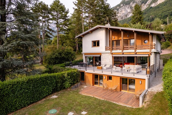 Una casa moderna de dos pisos con terraza de madera, ventanas amplias y montañas al fondo.
