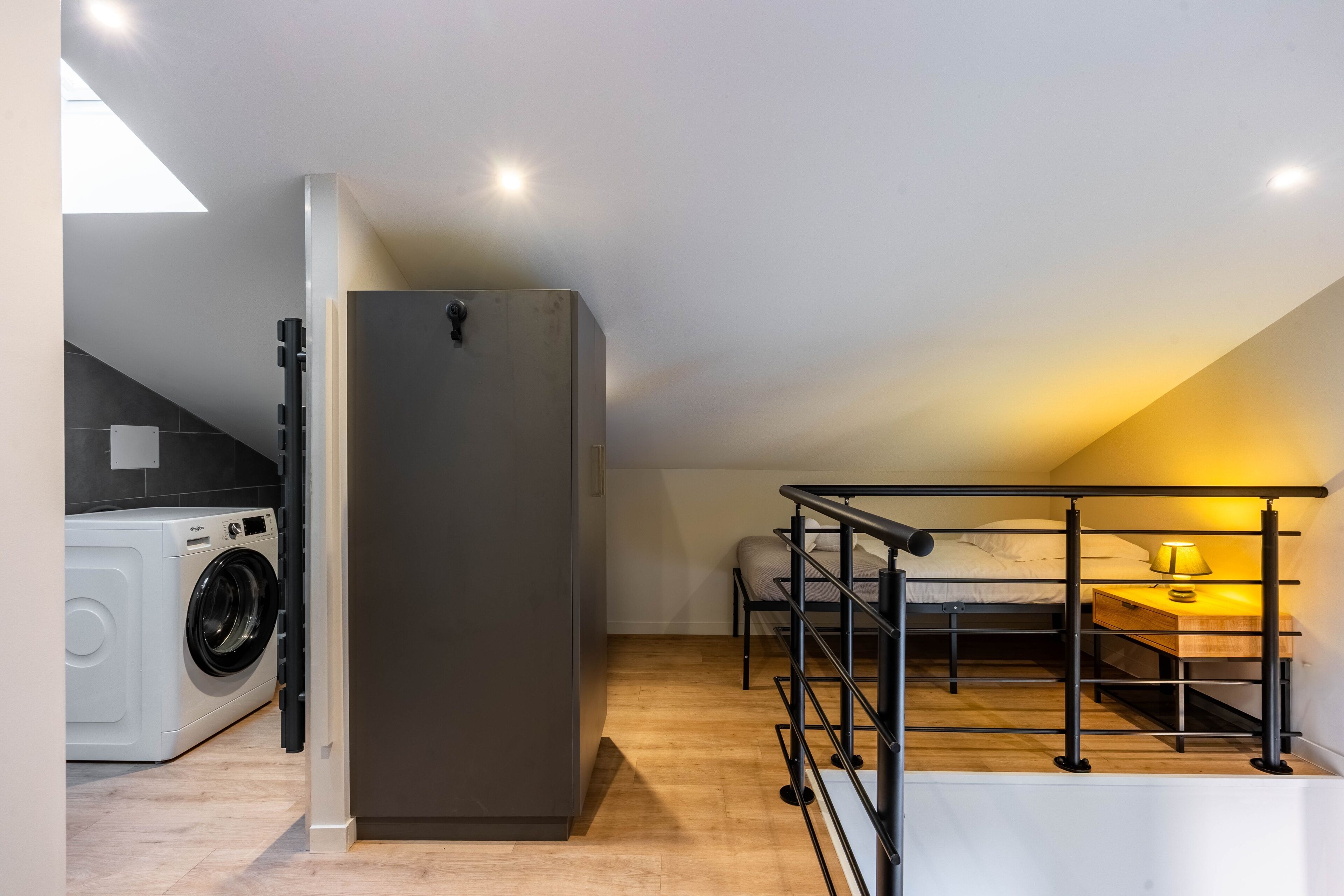 Eine kompakte, moderne Wohnung mit Küchenzeile, Waschmaschine, einem Bett mit Schreibtisch und Lampe sowie einem Balkon mit Geländer.