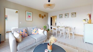 Ein modernes Wohnzimmer mit einem grauen Sofa, bunten Kissen, einem Esstisch mit Stühlen und einer weißen Kommode.