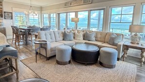 Ein geräumiges Wohnzimmer mit einer großen Eckcouch, Hocker, einem Essbereich mit Tisch und Stühlen sowie großen Fenstern mit Blick auf den Ozean.
