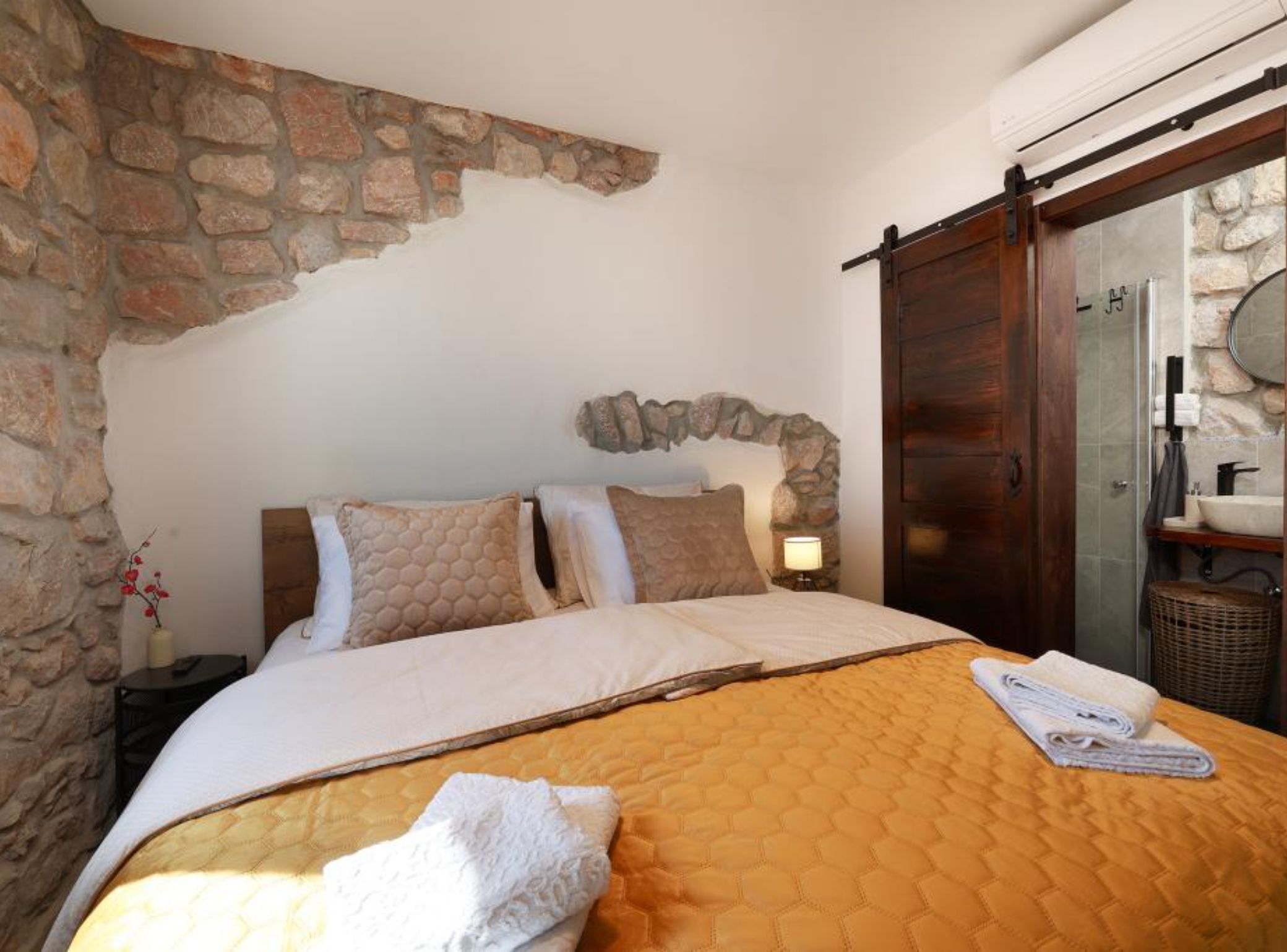 Un dormitorio con pared de piedra, cama con colcha, mesita de noche y baño con pared de piedra y puerta de madera.
