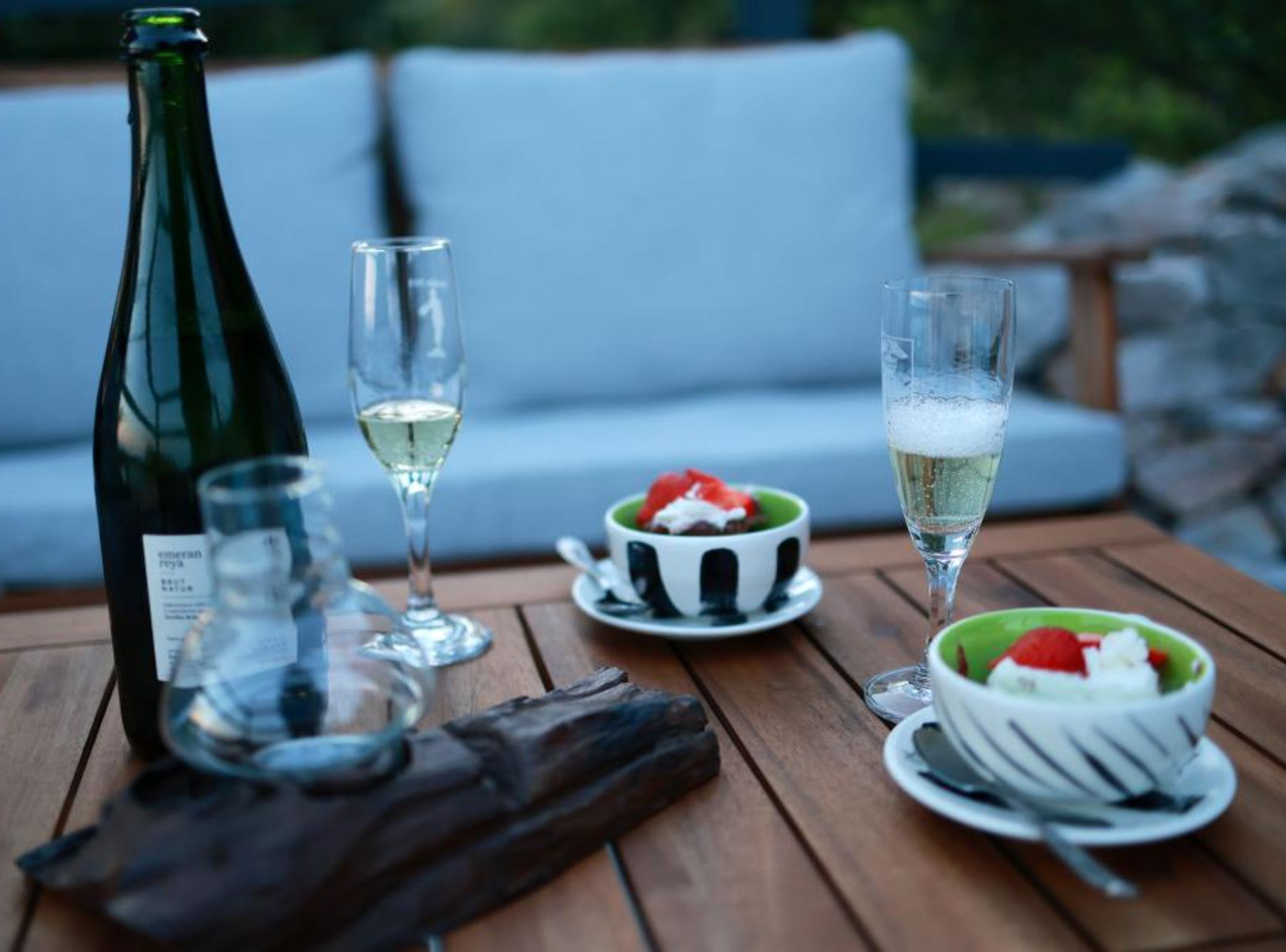 A table with a bottle of white wine, two glasses, and two bowls of dessert on a wooden table outdoors.