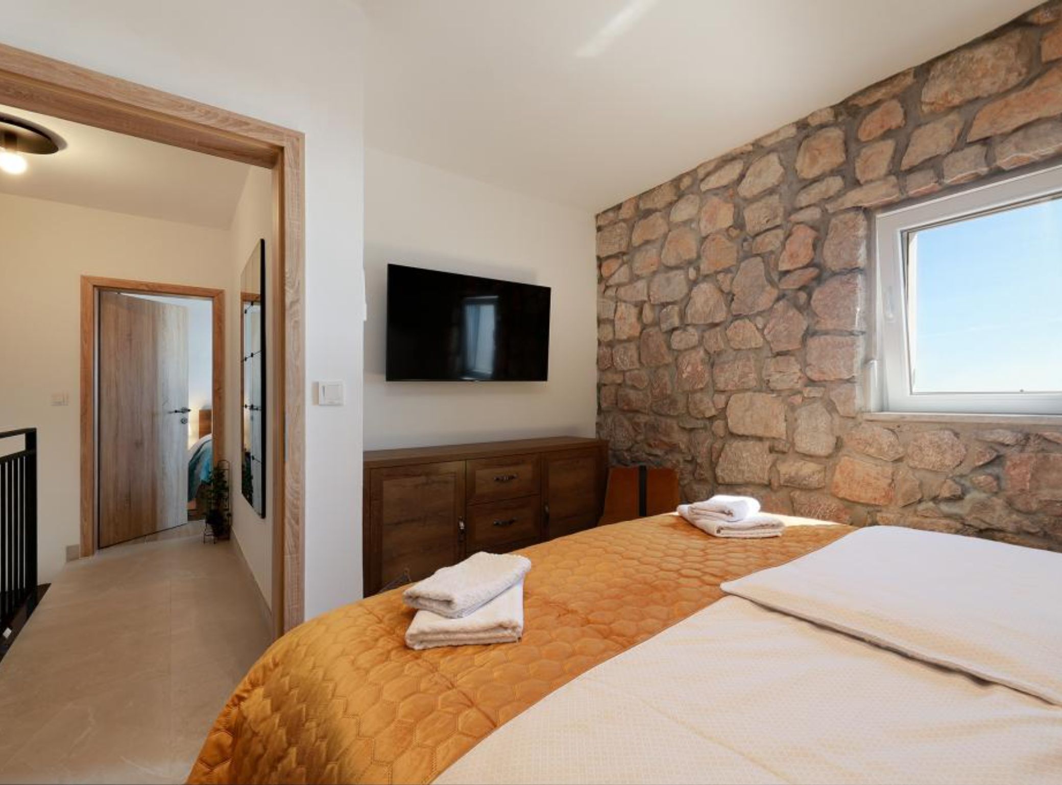 A bedroom with a stone wall, a flat-screen TV, a bed with two pillows, and a wooden dresser.