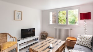 A living room with a TV, a wooden coffee table, wicker furniture, a red lamp, and a window with a view of greenery.