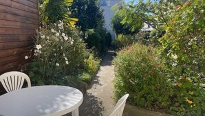 A narrow garden path leading to a wooden structure, surrounded by dense foliage and a white table with chairs.