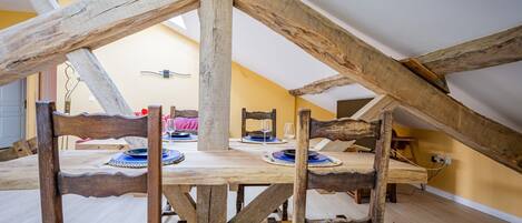 A dining area with a wooden table and chairs, a high ceiling with exposed beams, and a yellow wall in the background.