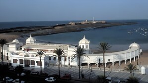 A coastal building with a curved facade, palm trees, and a view of the sea and a lighthouse in the distance. - Apartment 'La Bella Escondida' with Wi-Fi and Air Conditioning (Cadiz)