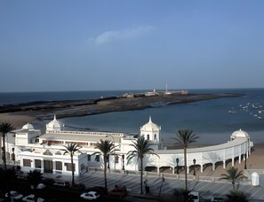 Ein Küstengebäude mit geschwungener Fassade, Palmen und Blick auf das Meer sowie einen Leuchtturm in der Ferne.