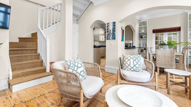 A spacious living area with a wooden staircase, wicker furniture, and a kitchen in the background. - Villa 17 on Graham (Cape Town)