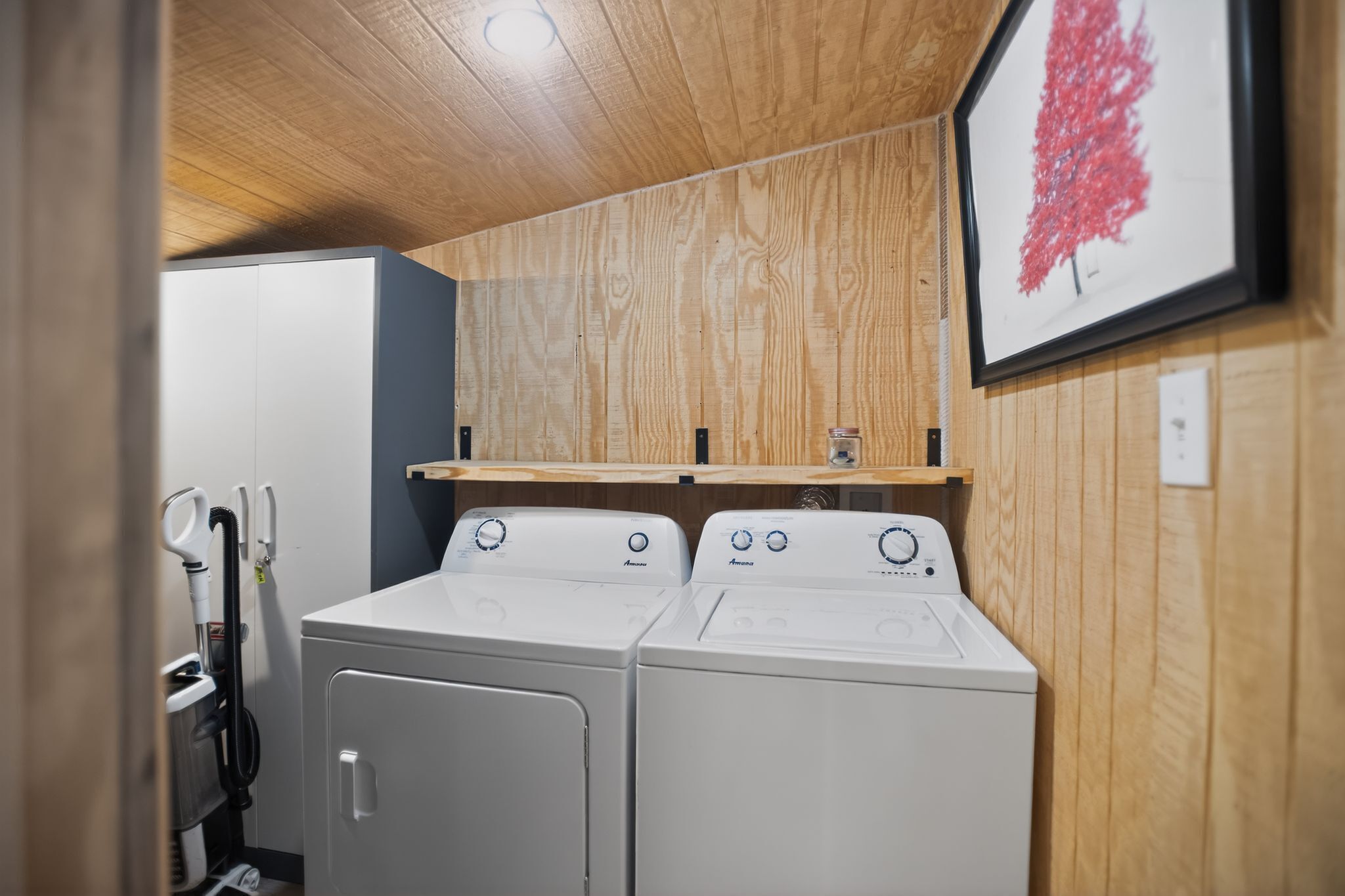 A laundry room with two white front-loading washers and dryers, wooden panel walls, and a shelf above the dryers.