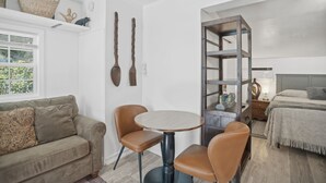 Une pièce de taille modeste comprenant un canapé, deux fauteuils, une table ronde, un lit, une étagère avec des objets décoratifs et une structure en forme d’échelle en bois.