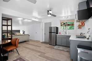 Cocina compacta con refrigerador, comedor con mesa y sillas, ventilador de techo y ventana con vista a la vegetación.