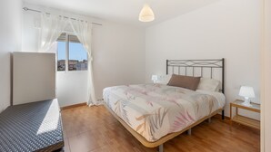 Et soveværelse med seng, et natbord, en lampe, et vindue med lette gardiner og udsigt til bygninger.