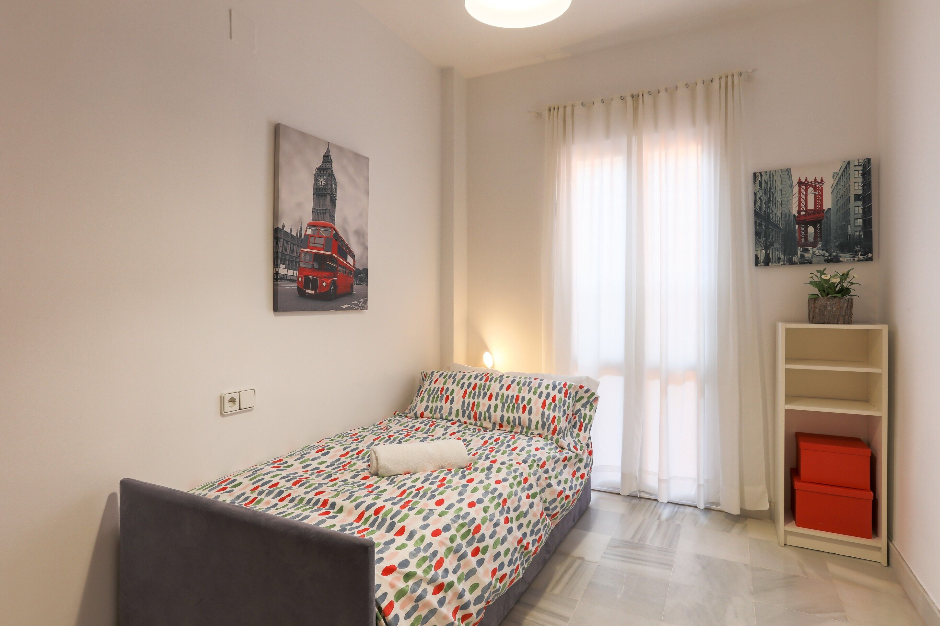 A neatly arranged bedroom with a bed, a chair, a shelf with storage boxes, and two wall-mounted pictures.
