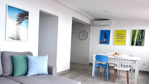 A modern living room with a grey sectional sofa, a dining area with a white table and blue chairs, and artwork on the walls. - Tartaga Penthouse (Adeje)