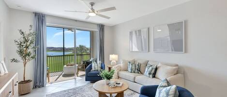 A modern living room with a large sliding glass door leading to a balcony with a view of a lake and greenery.