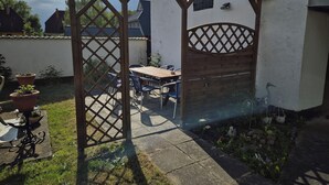 Una pérgola de madera con diseño de celosía, una mesa de patio con sillas y plantas en macetas.