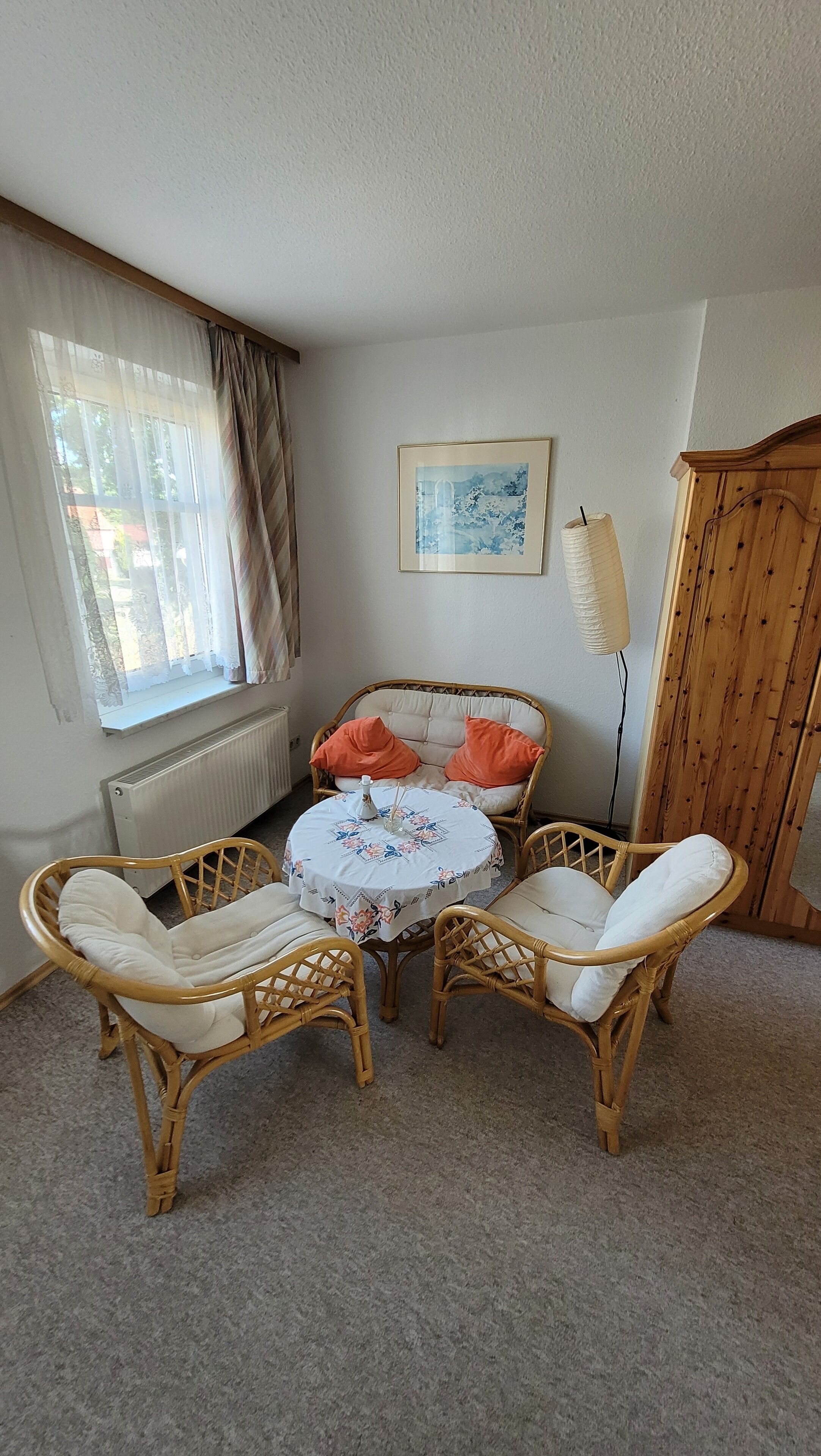A room with a round table, two wicker chairs, a radiator, a window with curtains, a wooden wardrobe, and a framed picture on the wall.
