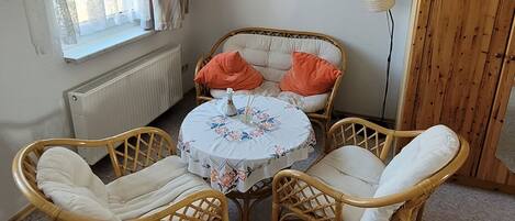 A room with a round table, two wicker chairs, a radiator, a window with curtains, a wooden wardrobe, and a framed picture on the wall.