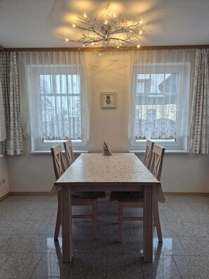 Un coin repas avec une table et des chaises en bois, deux fenêtres ornées de rideaux légers, un luminaire au plafond et un tableau représentant un ananas, encadré, accroché au mur.