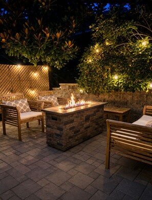 Een terras buiten met een vuurplaats, houten meubels en een stenen muur.