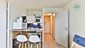 A compact kitchen with a microwave, coffee maker, and toaster. There are two white chairs and a small table. A door leads to another room. - Appartement 143 Résidence Bora Bora (Saint-Hilaire-de-Riez)