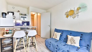 A compact living space with a kitchenette, a sofa with patterned cushions, and a wall art piece. - Appartement 143 Résidence Bora Bora (Saint-Hilaire-de-Riez)