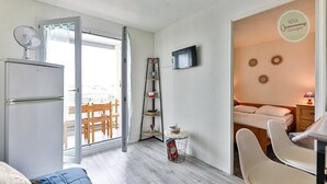 A compact hotel room with a bed, a desk, a chair, a TV, a small table, and a refrigerator. There is a balcony with a table and chairs visible through the open door. - Appartement 143 Résidence Bora Bora (Saint-Hilaire-de-Riez)
