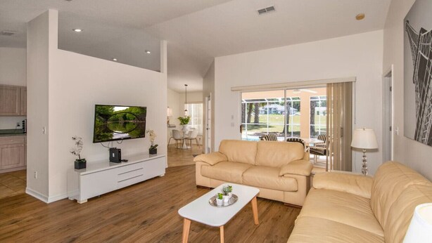 Amplia sala de estar con un sofá beige, un televisor de pantalla plana montado en la pared y un área de comedor visible a través de una puerta de cristal.