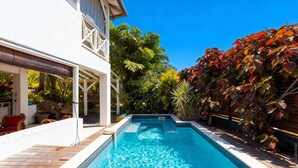 A swimming pool with a wooden deck, surrounded by lush greenery and a white building with a covered patio. - Granada (Le Diamant)